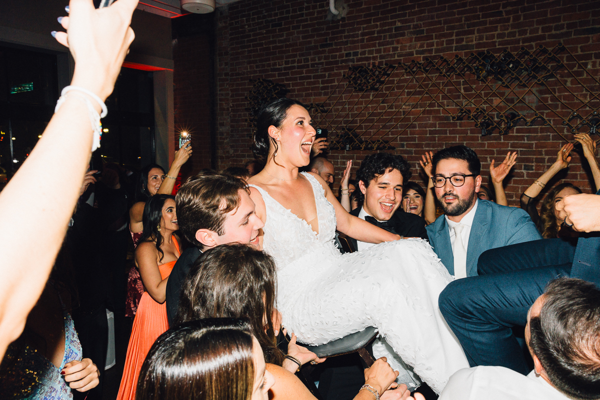 A bride joyfully lifted by friends during a lively dance at a wedding reception, surrounded by cheering guests.