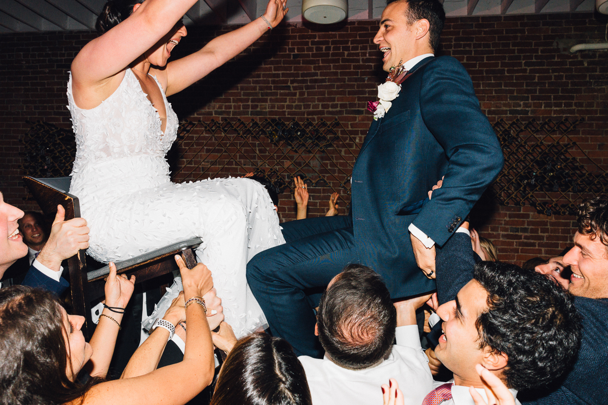 A joyful couple being lifted on chairs during a lively dance at their wedding reception, surrounded by enthusiastic friends and family.