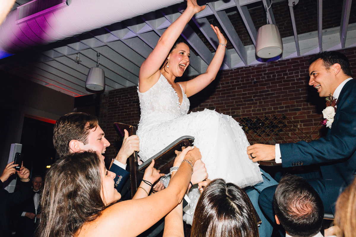 A joyful bride is lifted on a chair by guests during a wedding celebration, with smiles and excitement in the air.