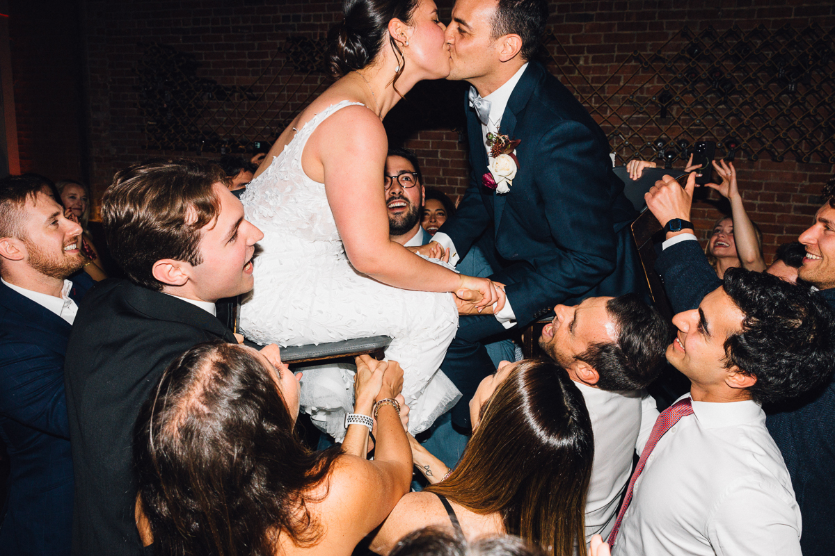 A bride and groom share a romantic kiss while being lifted on chairs by their friends during a lively celebration, surrounded by joyful guests.