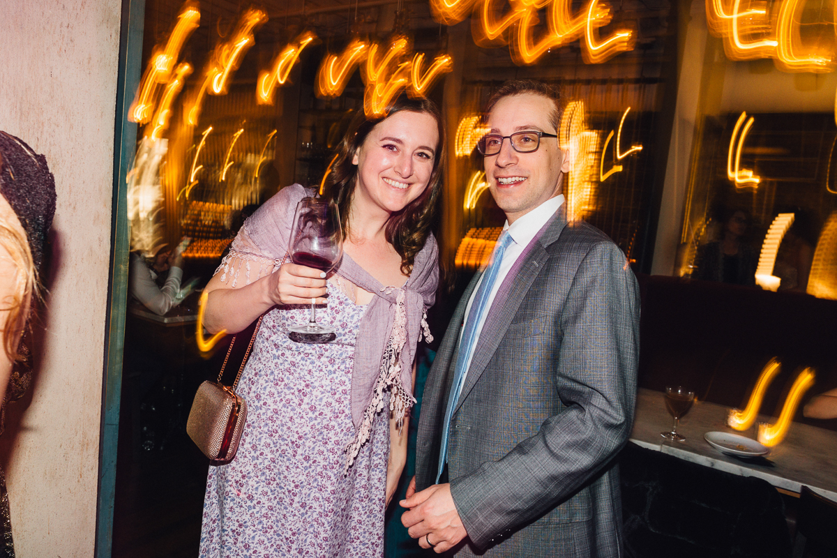 A woman holding a glass of red wine smiles beside a man in a suit, with vibrant background lights creating a festive atmosphere.