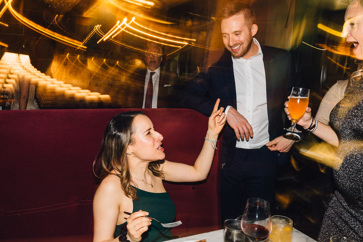 A woman gestures animatedly while seated at a dining table, surrounded by friends enjoying drinks in a lively restaurant atmosphere.