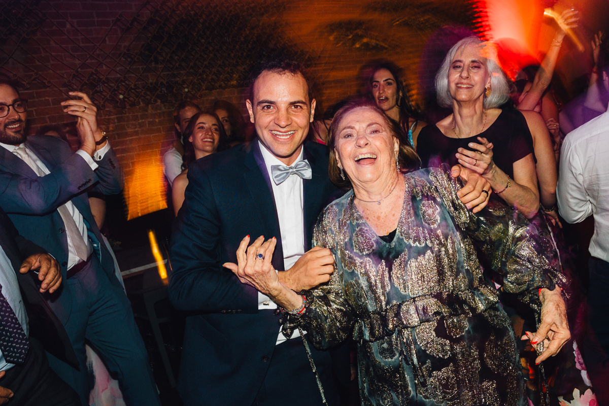 A man in a tuxedo and a woman in a patterned dress smiling and dancing together at a lively event, surrounded by people enjoying the moment.