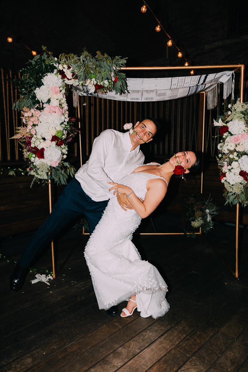 A playful couple dressed in wedding attire strikes a fun pose with roses in their mouths, surrounded by a floral arch.