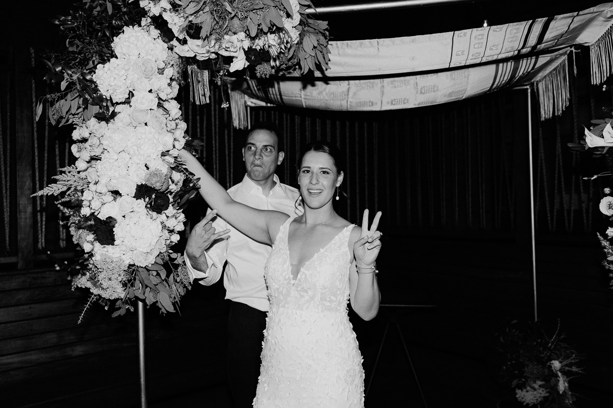 A bride playfully poses with a peace sign while standing under a floral arch, as a man makes a humorous expression in the background.