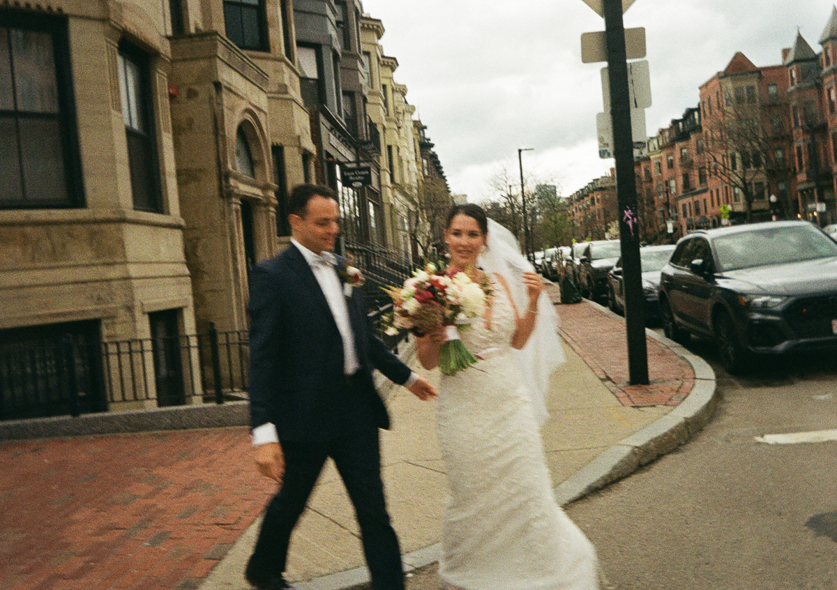 A joyful couple walking down a street, with the bride holding a bouquet and wearing a wedding dress while the groom supports her, amidst urban architecture and parked cars.