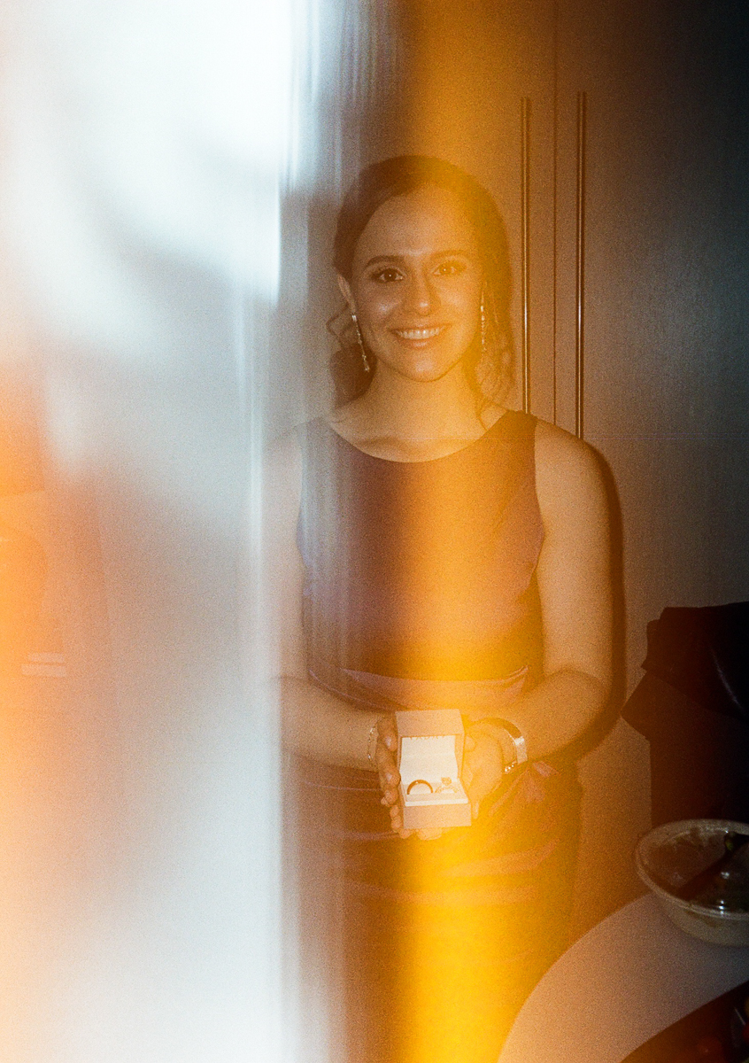 A smiling woman in a formal dress holds an open ring box, displaying elegant rings, with soft lighting and a blurred background.