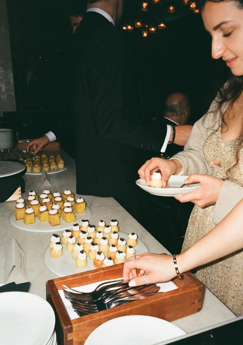 A woman in a sparkling outfit carefully selects a dessert from a table filled with various sweet bites, surrounded by elegantly dressed guests.
