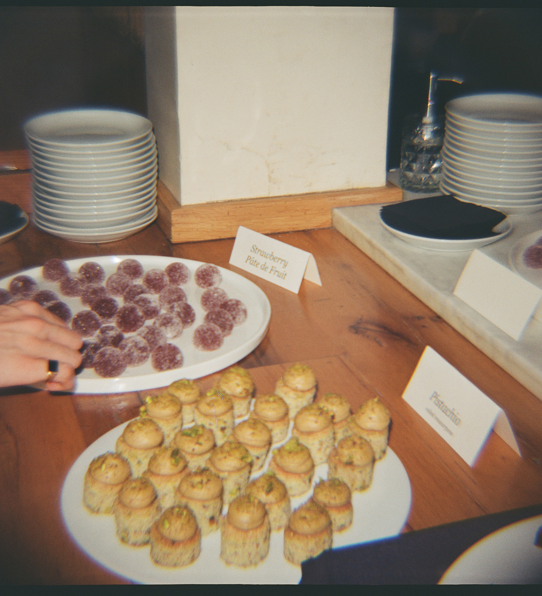 An assortment of desserts featuring strawberry pâte de fruit and pistachio treats on a wooden table, with stacked plates in the background.