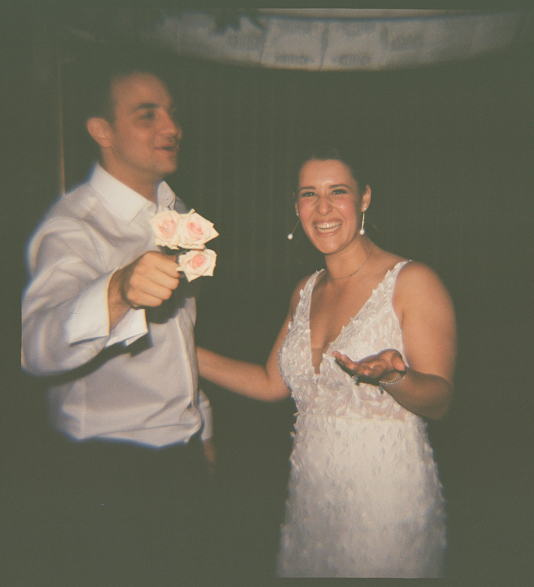 A bride and groom share a joyful moment, with the groom holding a bouquet of pink roses and the bride smiling brightly in her elegant wedding dress.