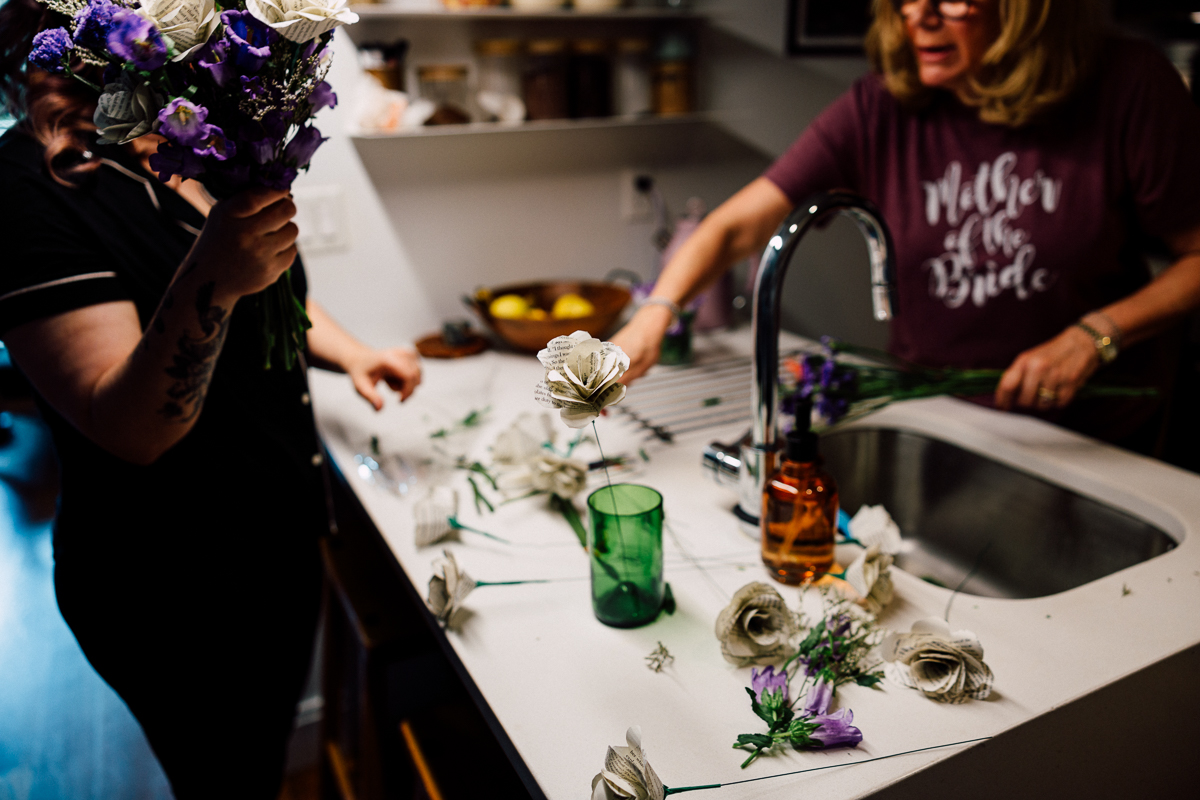 Two women crafting floral arrangements in a kitchen, with decorative flowers scattered on the countertop and a "Mother of the Bride" shirt visible.