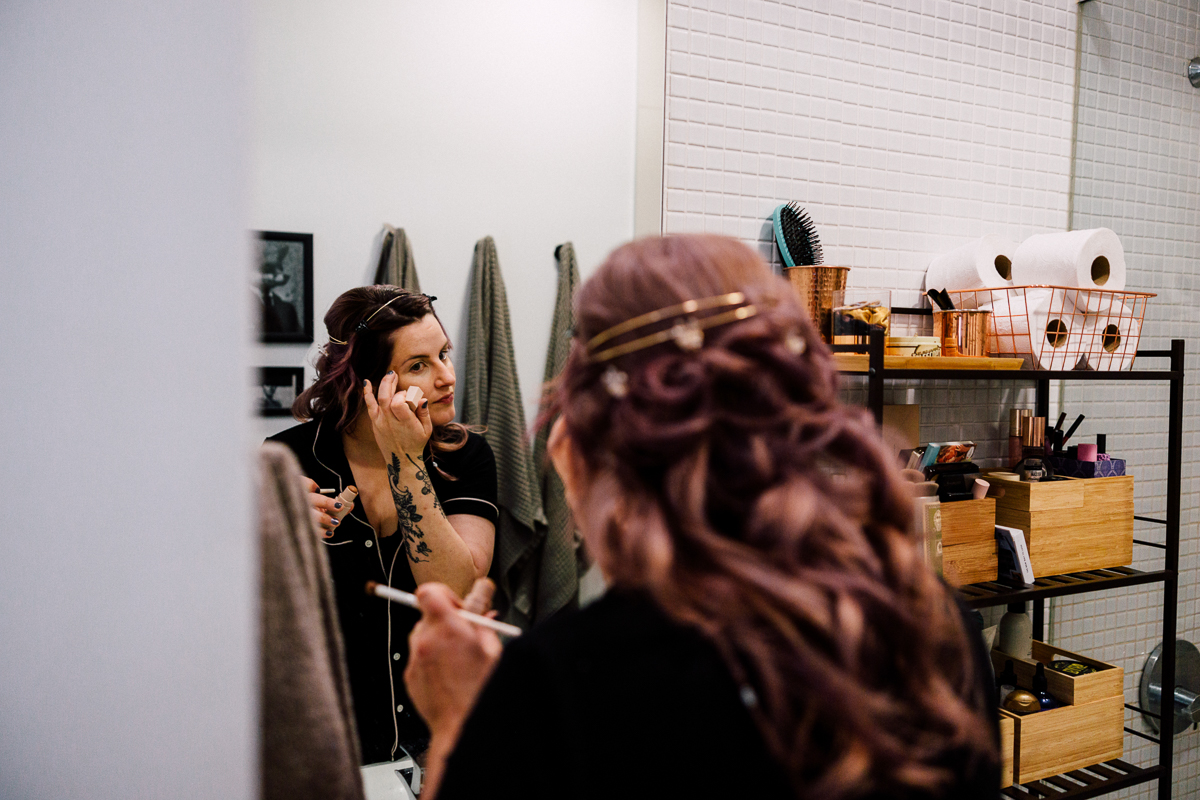 A woman with a tattoo applying makeup in front of a bathroom mirror, surrounded by beauty products and towels.