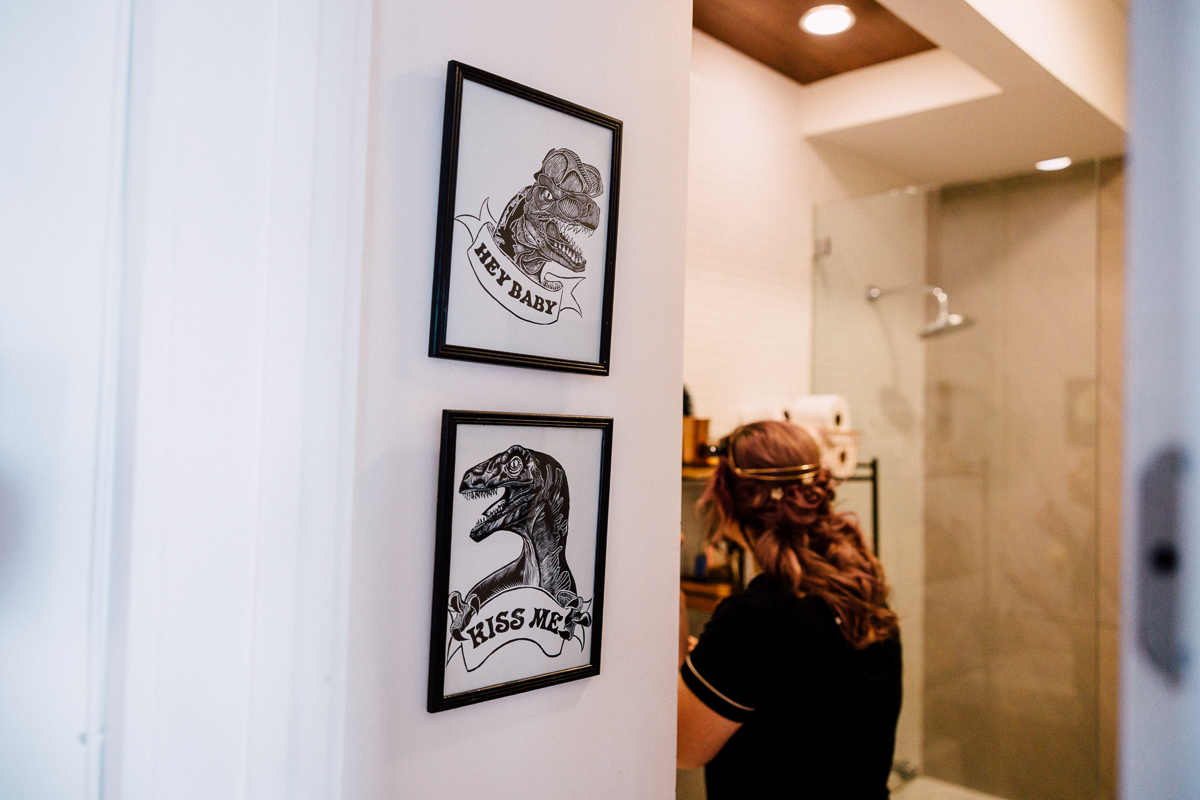 A woman with a braided hairstyle stands in a bathroom near two framed prints featuring dinosaur illustrations with playful captions.