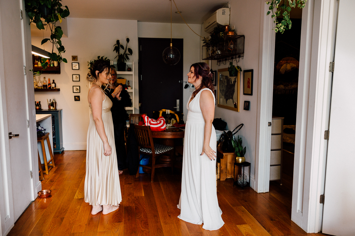 Two women in elegant dresses converse in a warmly lit room filled with plants and decor, as they prepare for a special occasion.