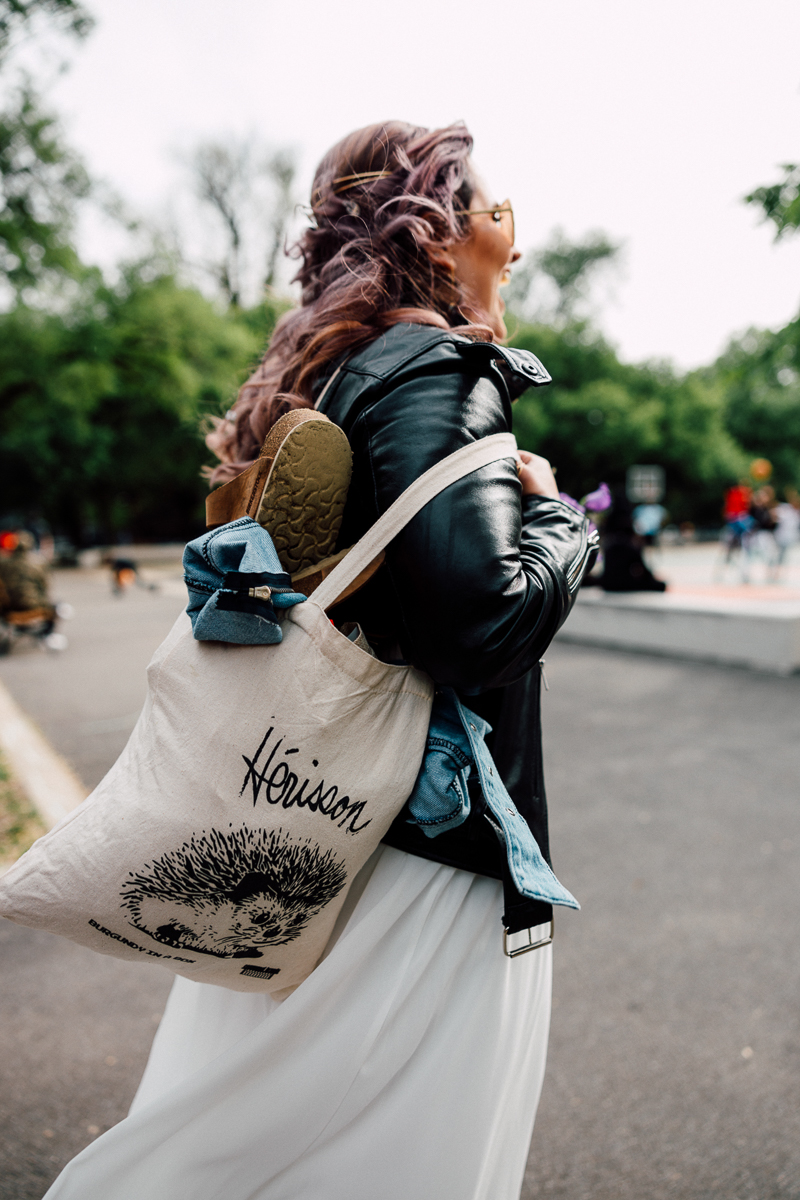 A woman with purple hair wearing a leather jacket and white skirt carries a tote bag featuring a hedgehog design, with a pair of sandals and a denim jacket peeking out.