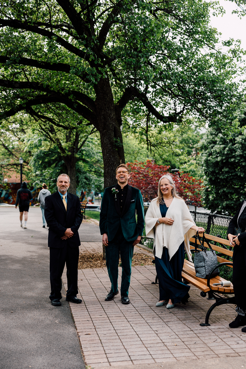 Three smiling individuals dressed in formal attire stand together in a park setting, surrounded by lush greenery and a pathway.