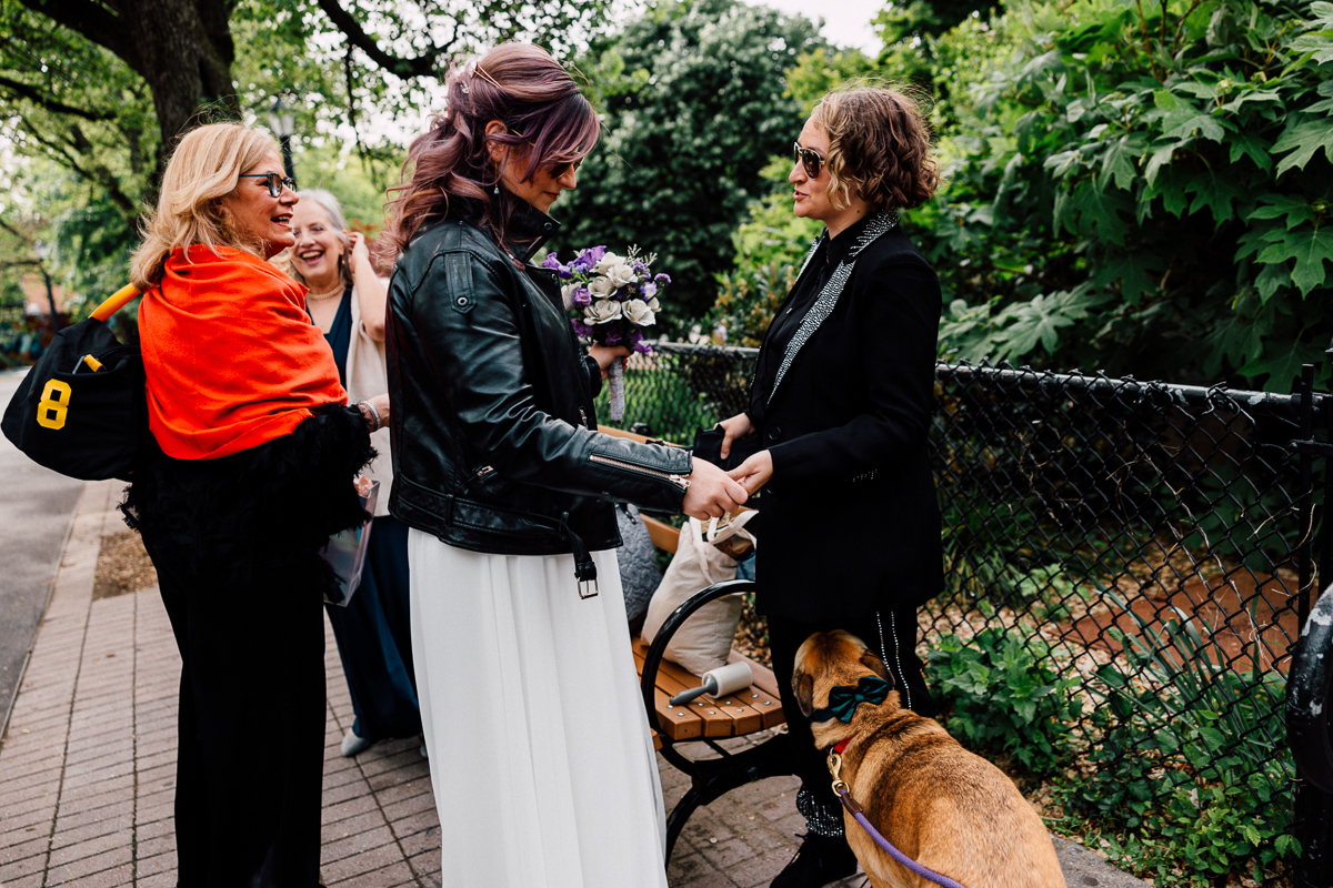 A group of friends gathers in a park, with one woman holding a bouquet while engaging with another, surrounded by greenery and a dog.