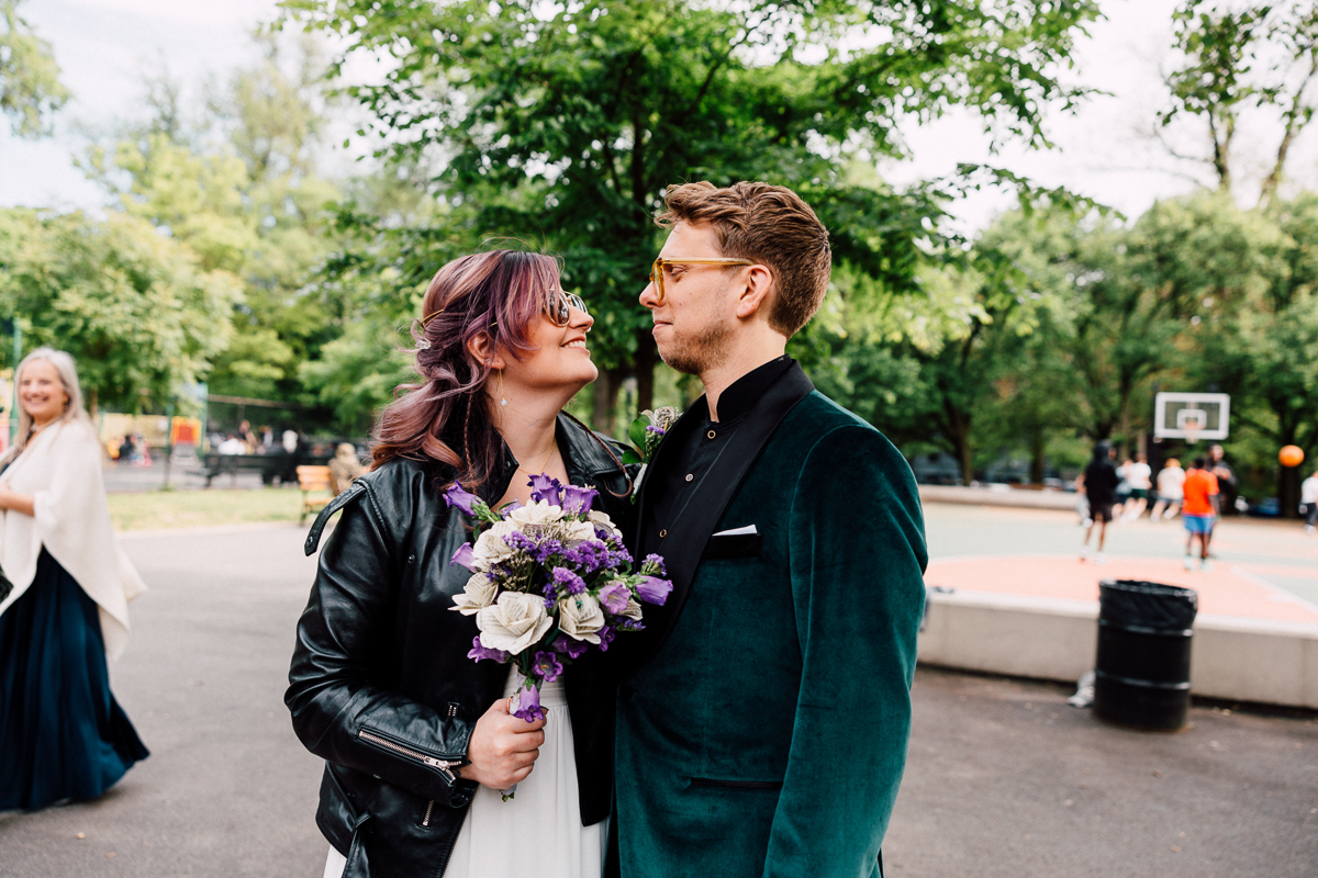 A couple sharing a joyful moment in a park, the woman wearing a black leather jacket and holding a bouquet, while the man dons a stylish green suit, with a basketball court visible in the background.