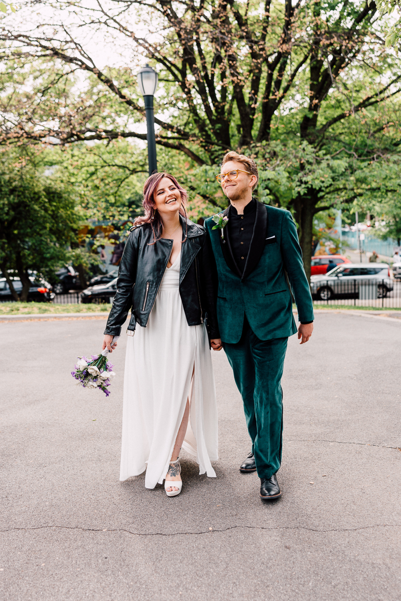 A couple walks hand in hand, smiling, with the woman in a white dress and leather jacket, and the man in a green velvet suit, surrounded by greenery.