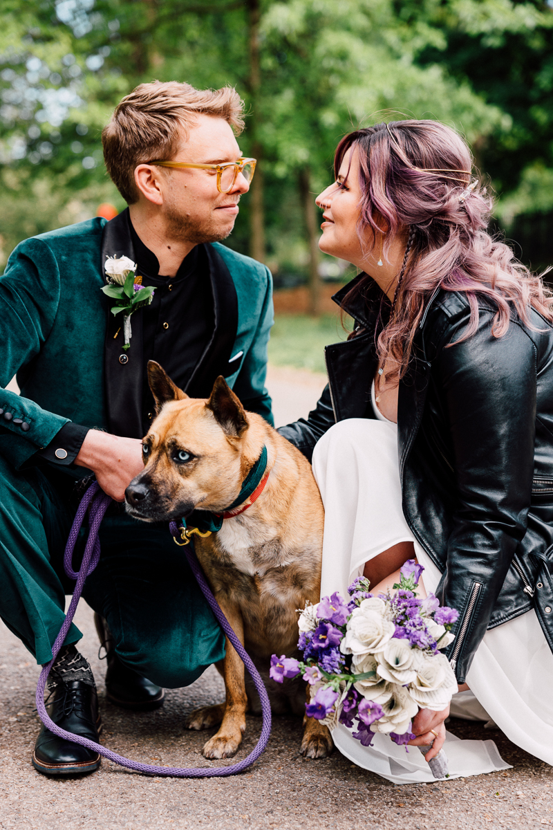A joyful couple kneels in a park, lovingly gazing at each other while holding a dog and a bouquet of flowers, embodying a modern wedding celebration.