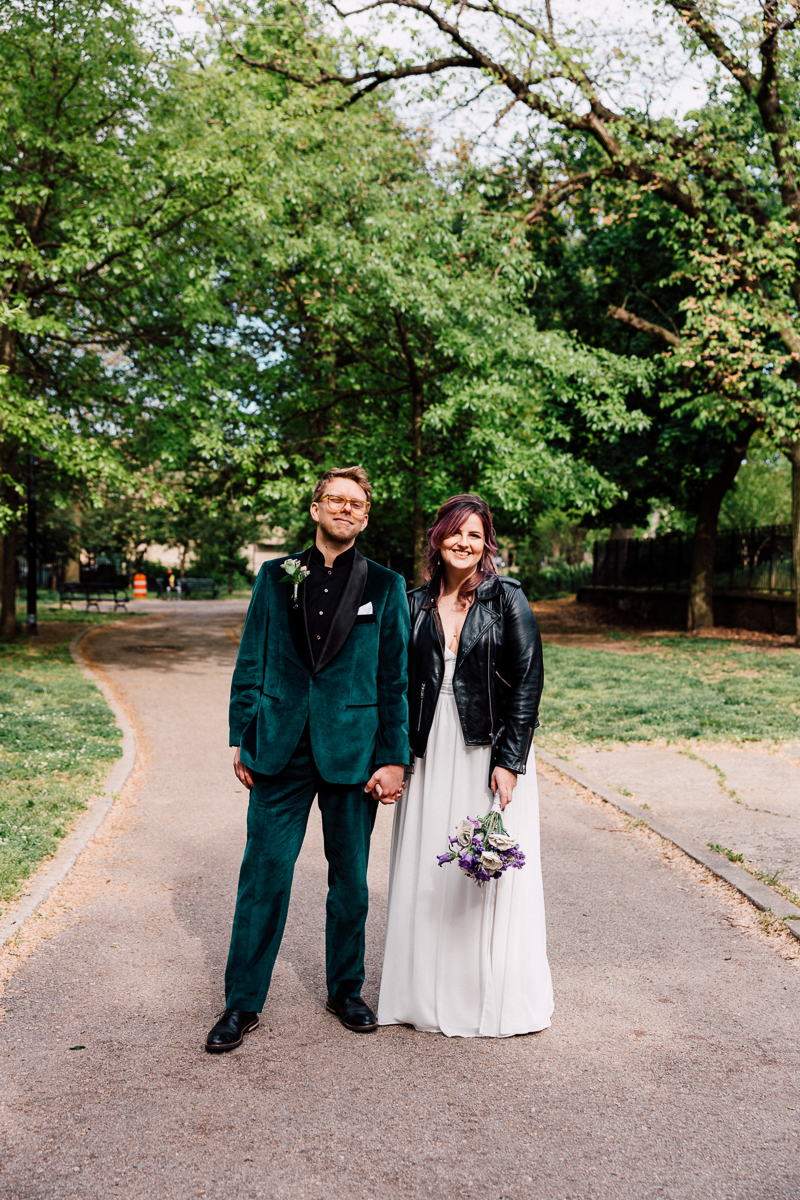 A happy couple stands hand-in-hand on a park pathway, surrounded by lush greenery, with the groom in a vibrant green suit and the bride in a white dress and leather jacket holding a bouquet.