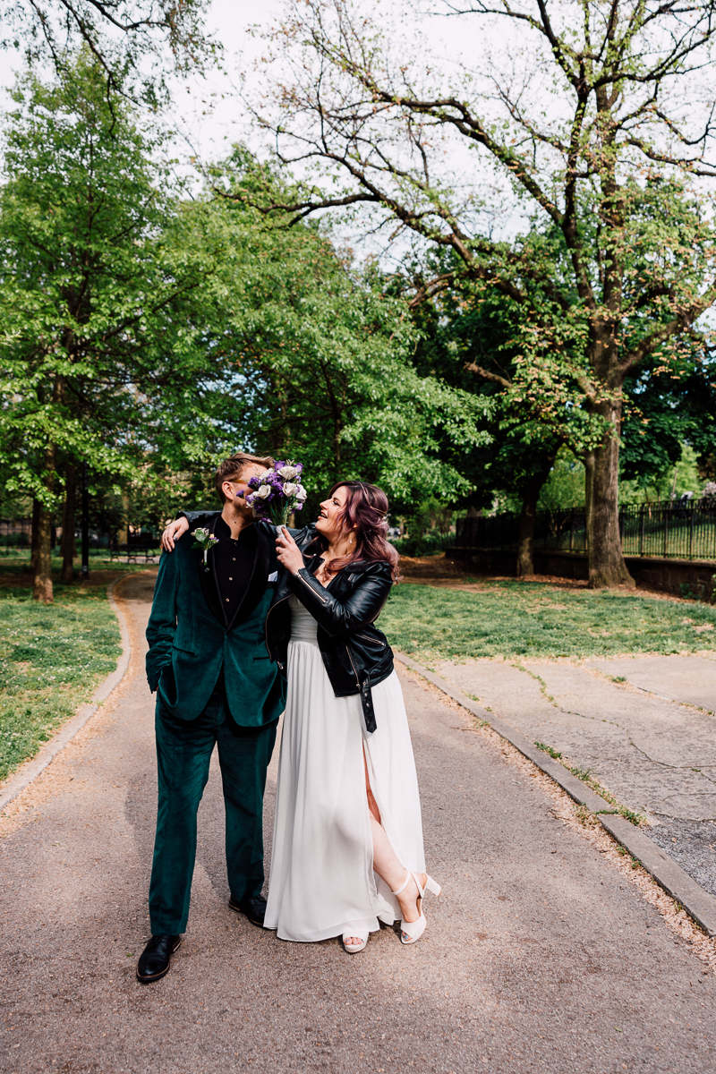 A couple shares a playful moment in a park, with the woman playfully holding a bouquet of flowers near the man's face. The scene is framed by lush greenery and a winding path.