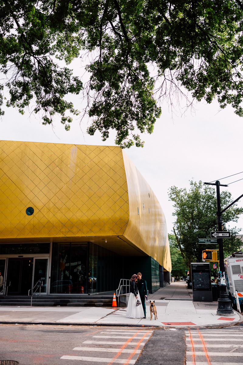A couple in wedding attire stands with a dog outside a striking yellow modern building, surrounded by trees and city elements.