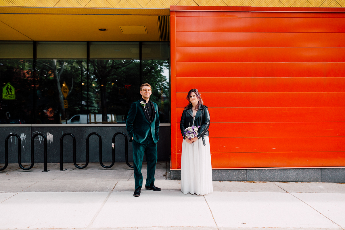A bride in a white dress and leather jacket stands with a groom in a green suit in front of an orange wall, showcasing a vibrant urban setting.