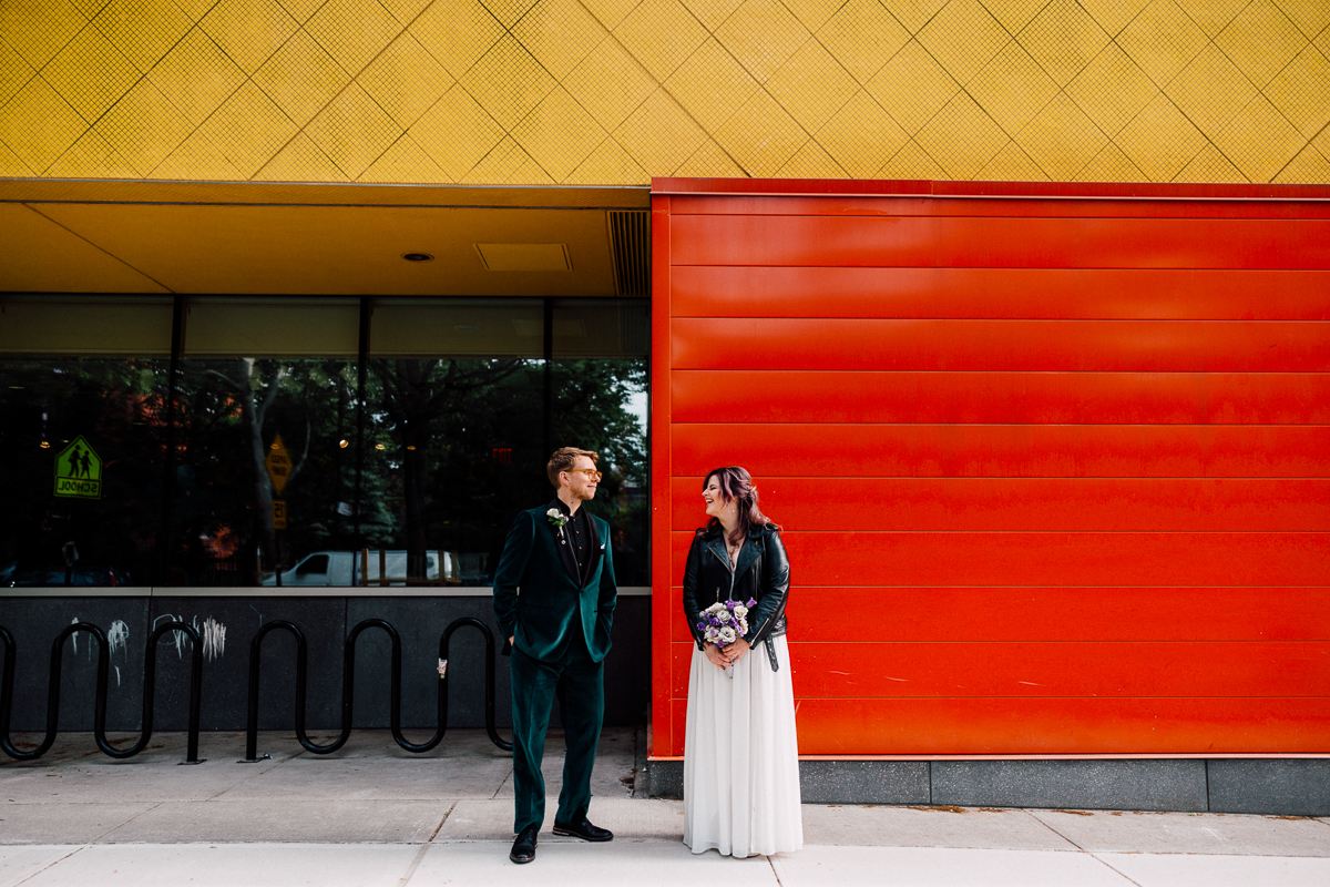 A couple poses playfully outdoors, with vibrant yellow and red walls creating a striking backdrop for their wedding attire.