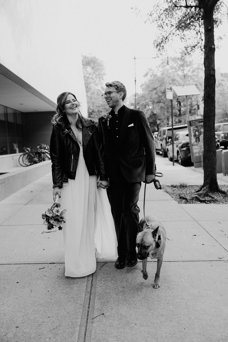 A couple dressed in wedding attire, walking hand in hand with a dog, surrounded by an urban setting.