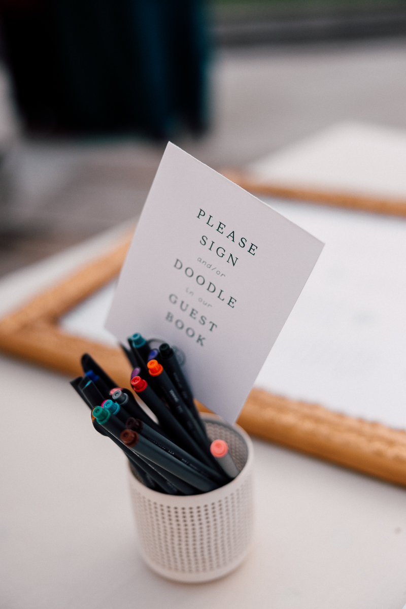A white sign instructing guests to sign and doodle in the guest book, accompanied by a collection of colorful pens in a holder.