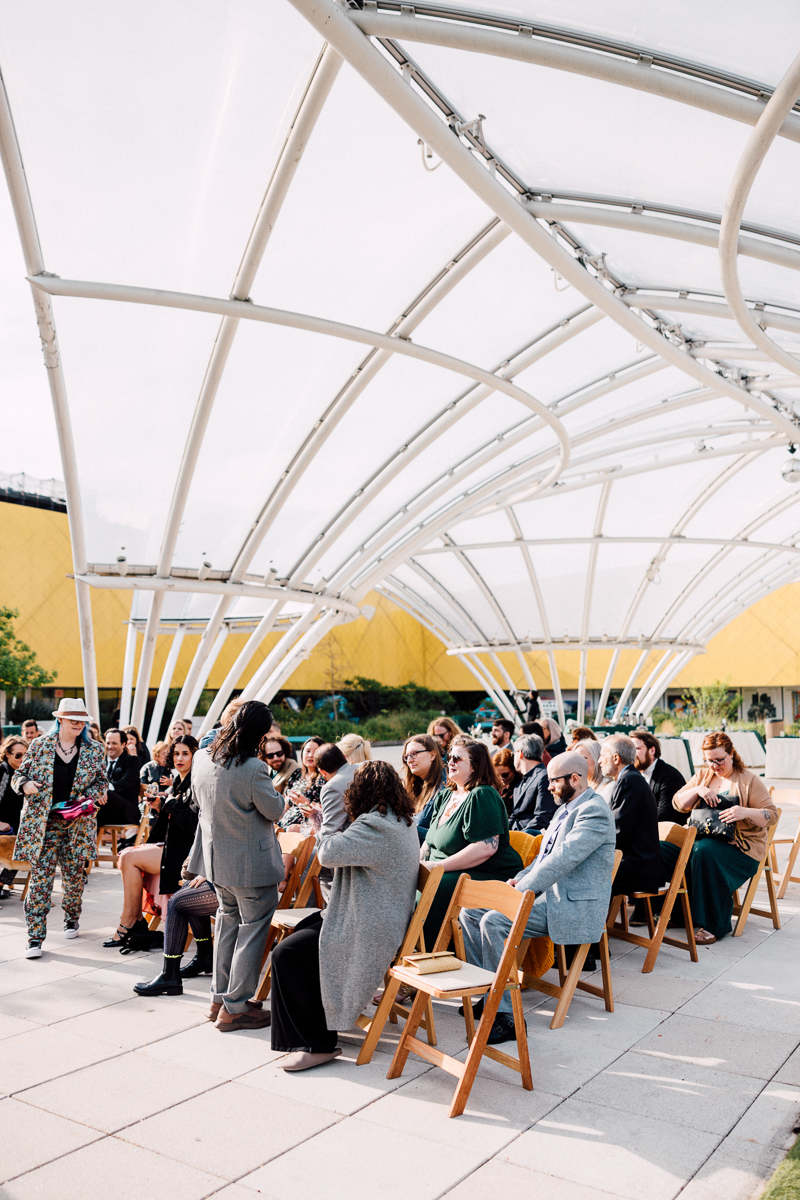 A diverse group of seated guests in an outdoor setting beneath a curved transparent structure, with lively interactions and a vibrant atmosphere.