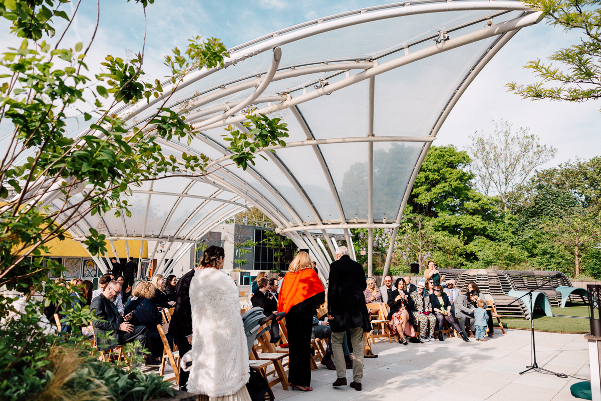 A spacious outdoor venue featuring guests seated under a modern transparent canopy, surrounded by greenery and open skies.