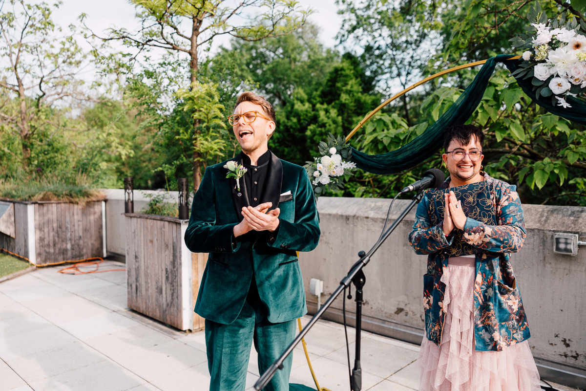 Two people stand outdoors at a decorated microphone during a Brooklyn Childrens Museum Wedding. One, in a green velvet suit with a boutonnière, smiles holding a phone; the other, in a floral jacket and pink skirt, stands with hands together, smiling warmly.
