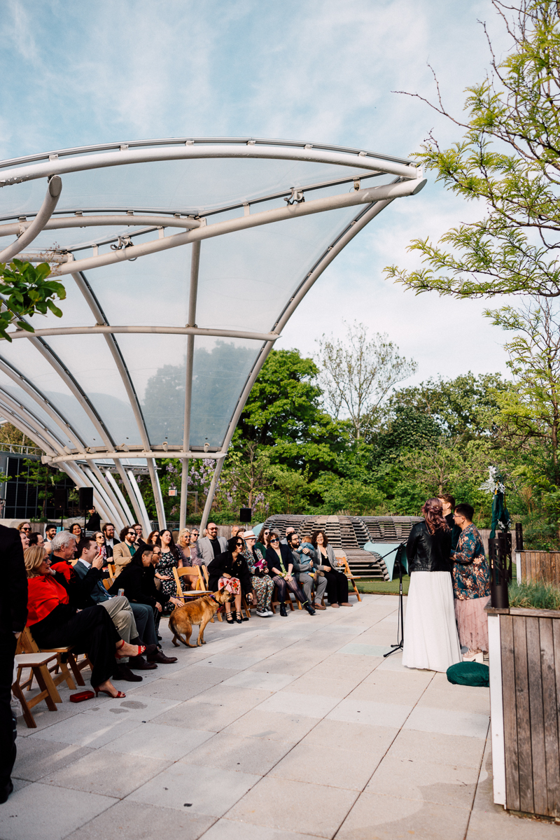 A wedding ceremony takes place outdoors under a modern, curved canopy. Guests sit on benches, some with cameras, as two people stand at the altar with an officiant. A dog sits among guests—an atmosphere akin to a Brooklyn Childrens Museum Wedding.