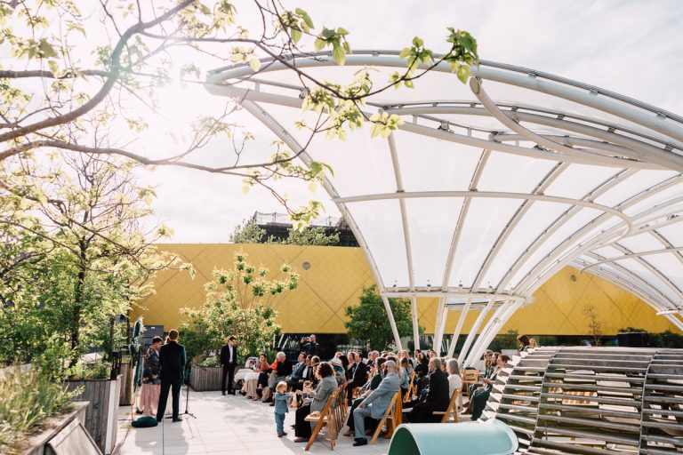 An outdoor Brooklyn Childrens Museum Wedding unfolds beneath a modern, curved glass canopy. Guests sit on wooden chairs amid lush greenery and sunlight, with the iconic yellow museum building in the background.