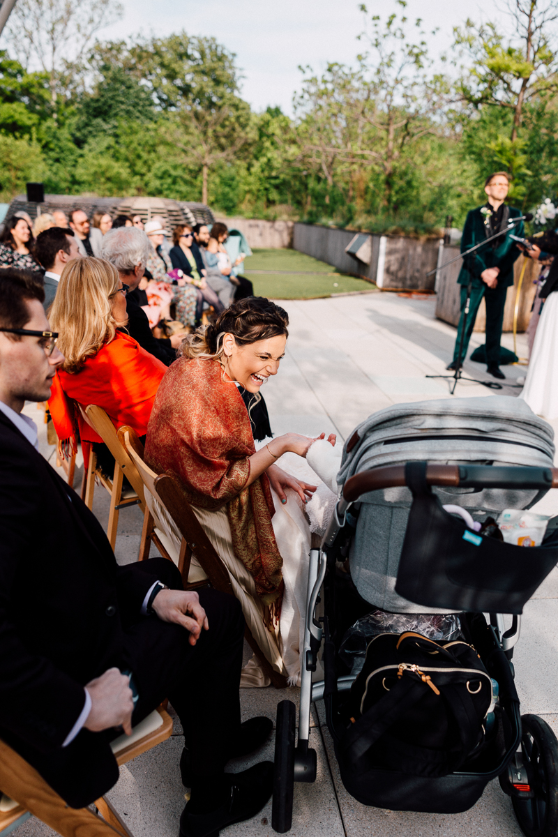 At a Brooklyn Childrens Museum Wedding, a woman in a colorful shawl smiles at a baby in a stroller while sitting among guests. Rows of attendees face a speaker at the front, surrounded by lush trees and greenery.