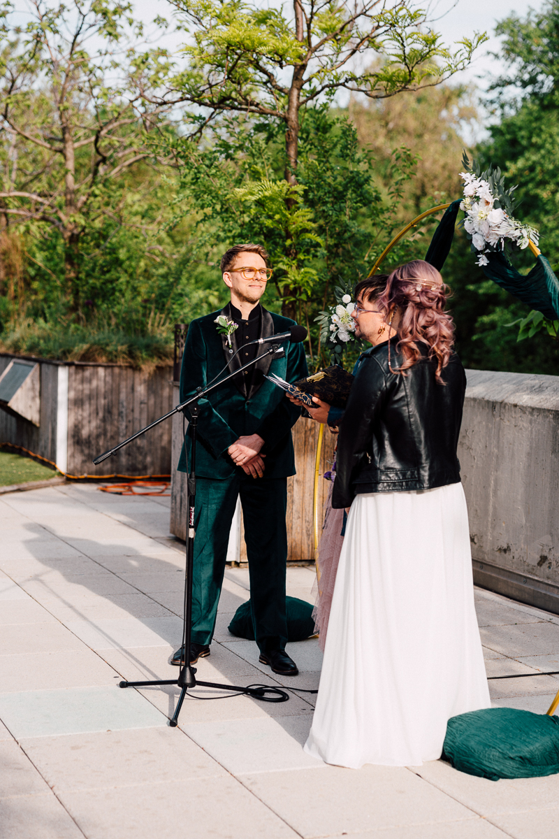 A couple stands facing each other at an outdoor Brooklyn Children's Museum wedding. The groom wears a dark green suit, and the bride, in a leather jacket over her white dress, stands by a microphone. They are surrounded by lush greenery.