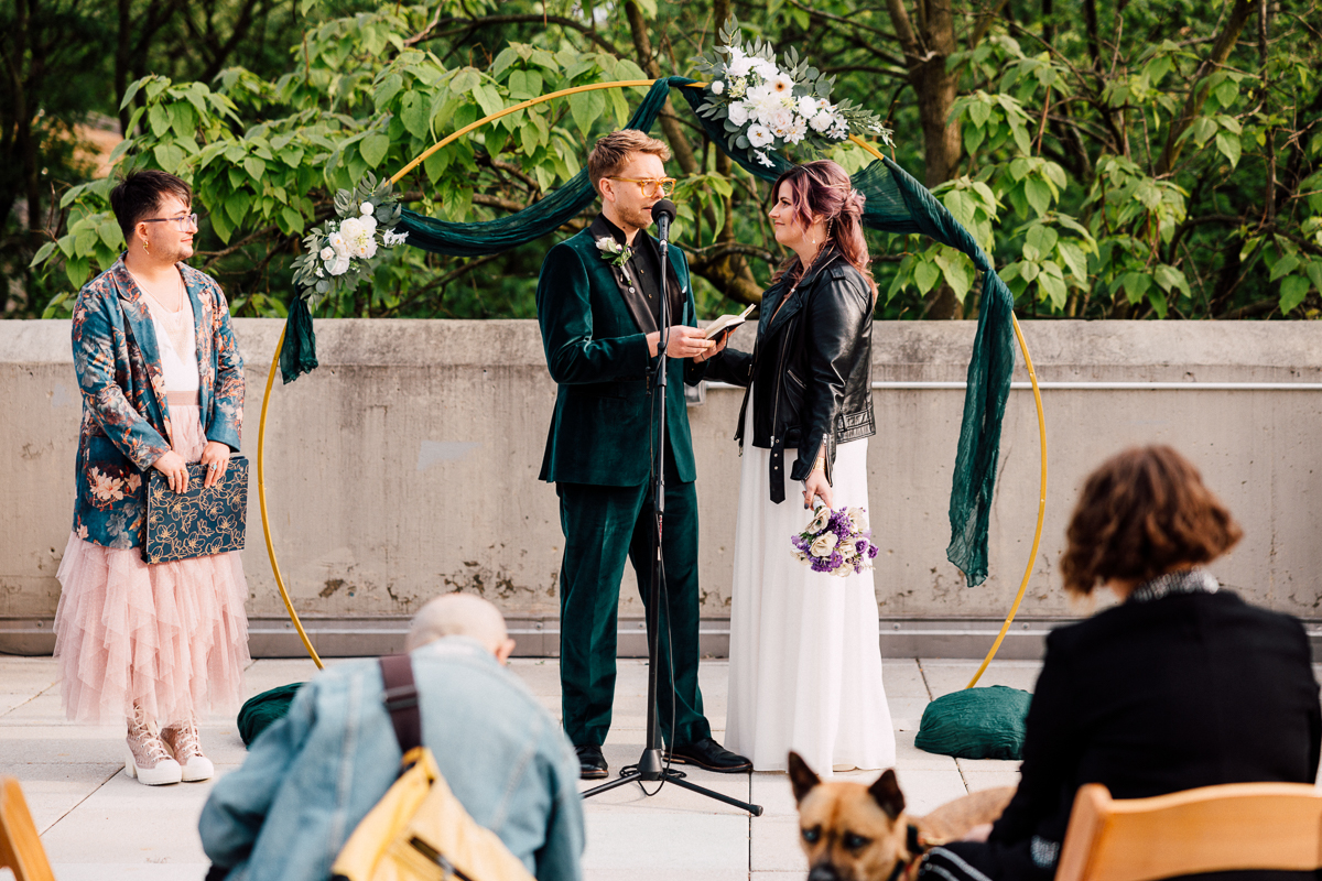 A couple stands under a decorated gold arch during their outdoor Brooklyn Childrens Museum Wedding ceremony. The groom reads vows from paper, while the bride in a leather jacket holds a bouquet. An officiant and guests look on.