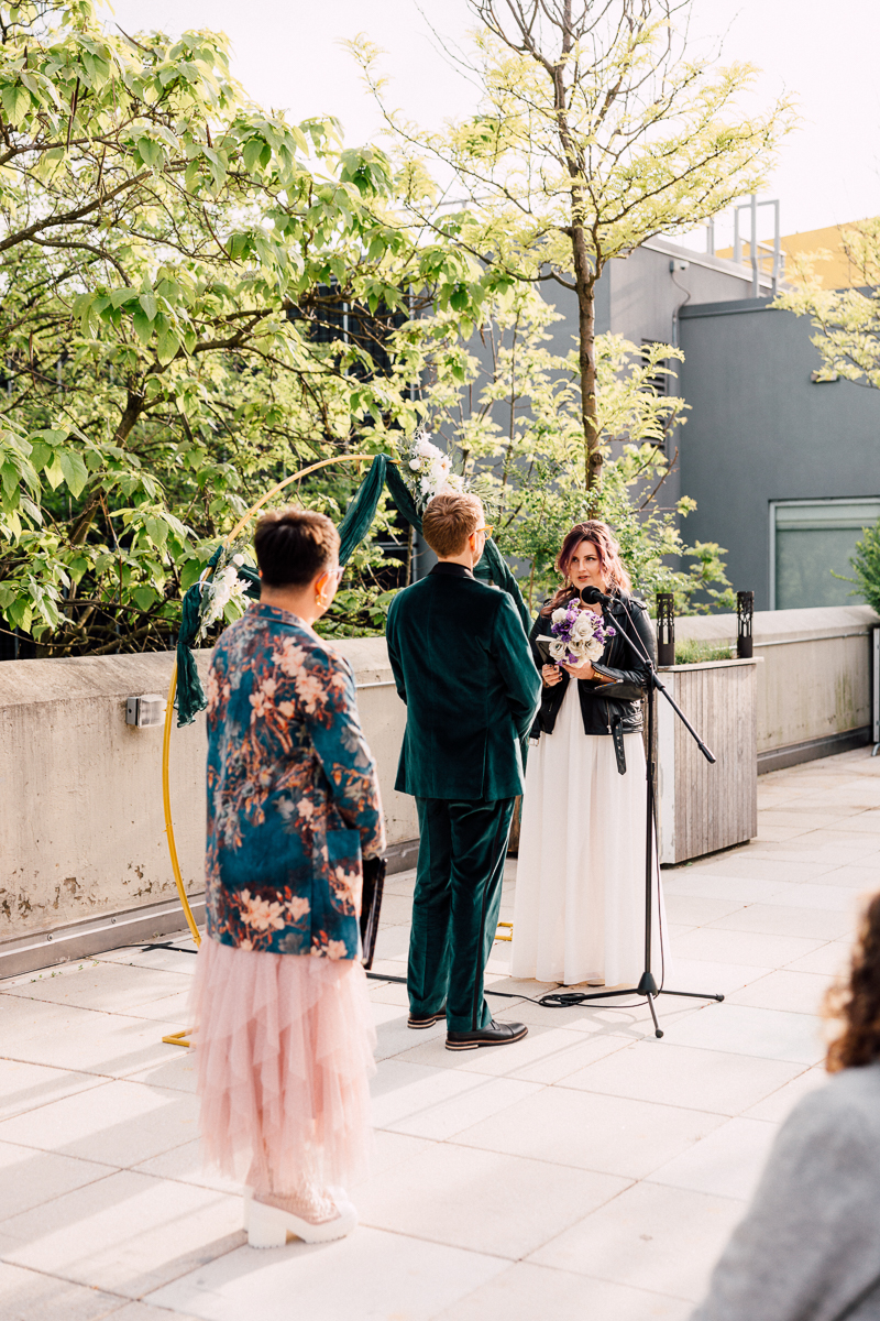 A couple stands at a microphone during an outdoor Brooklyn Children’s Museum Wedding ceremony, with one holding a bouquet. Nearby, a guest in a floral jacket and pink tulle skirt stands as green trees and the modern museum building create a vibrant backdrop.