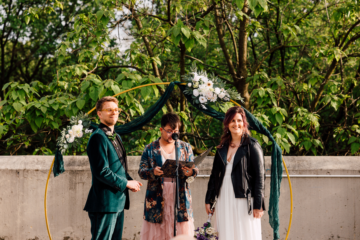 A couple stands facing an officiant under a circular floral arch outdoors at their Brooklyn Children’s Museum wedding. The groom wears a teal suit and bow tie, while the bride pairs her white dress with a black jacket amid lush green trees.