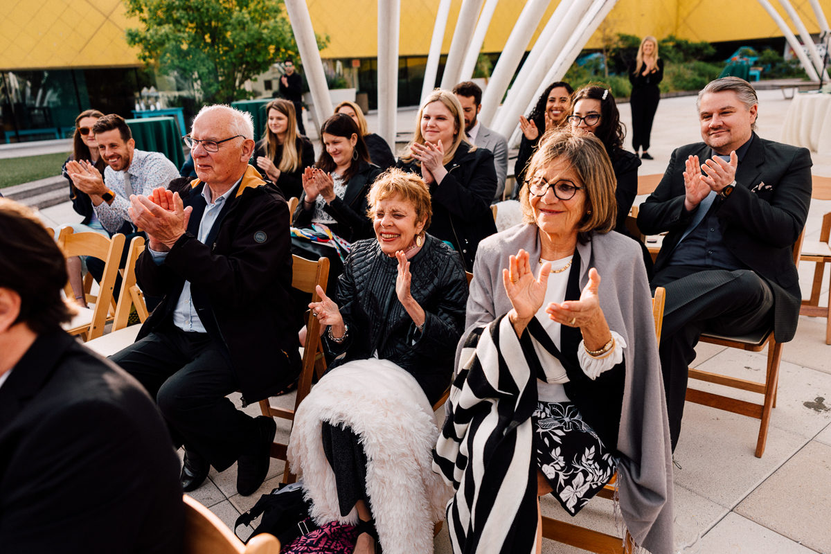 A group of people sitting outdoors on wooden chairs, smiling and applauding at a Brooklyn Childrens Museum Wedding. The setting appears festive with bright sunlight, greenery, and a modern yellow building in the background.