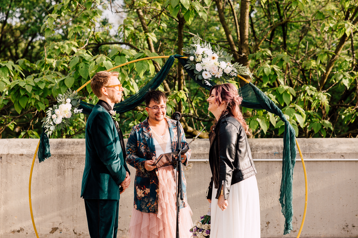 A couple stands facing each other during an outdoor Brooklyn Children’s Museum Wedding ceremony. An officiant reads from a book between them under a circular floral arch, with greenery and trees in the background.