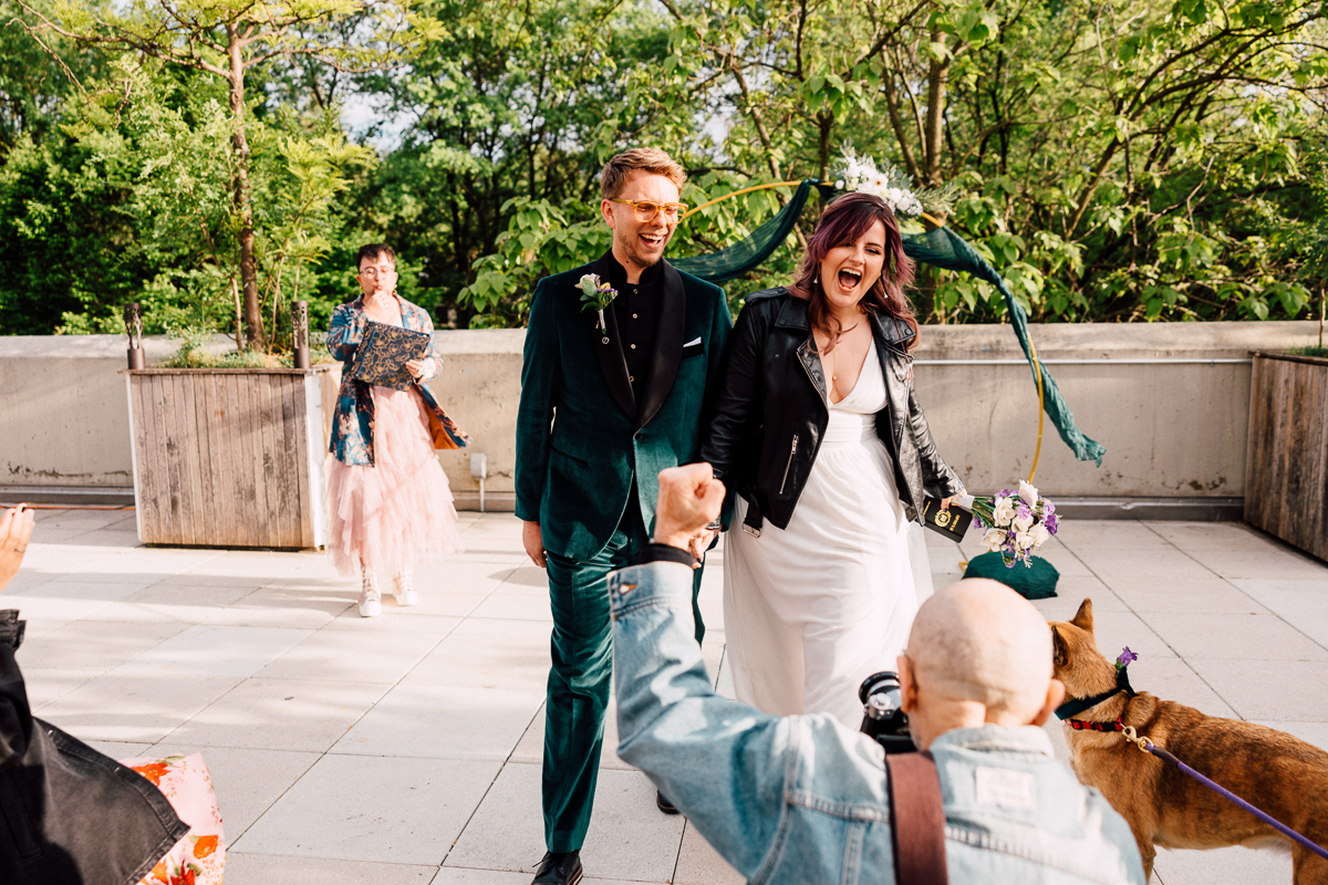 A joyful couple walks outdoors after their Brooklyn Childrens Museum Wedding; the groom in a teal suit and orange glasses, the bride in a white dress with a black jacket and green ribbons. Friends, family, and a dog greet them amid lush greenery as they smile and laugh.