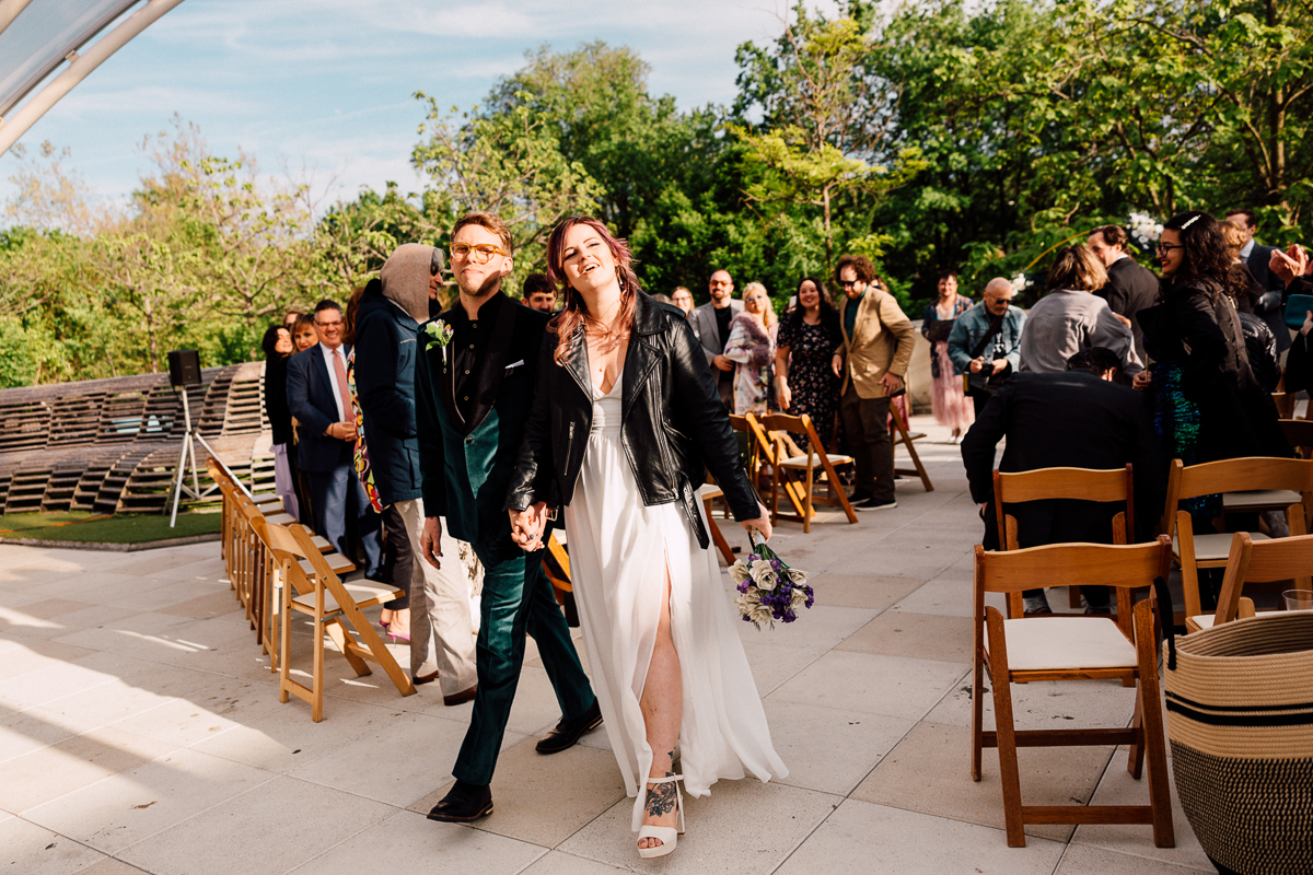 A couple walks hand in hand, smiling broadly as they leave a wedding ceremony surrounded by guests and greenery.
