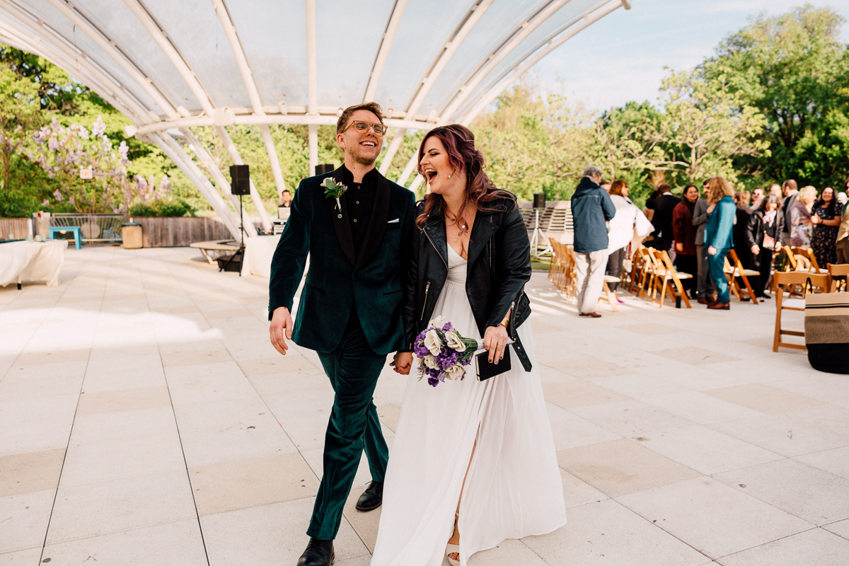 A happy couple walks hand in hand with smiles, the groom in a green suit and the bride in a white dress with a leather jacket, surrounded by a festive outdoor setting.