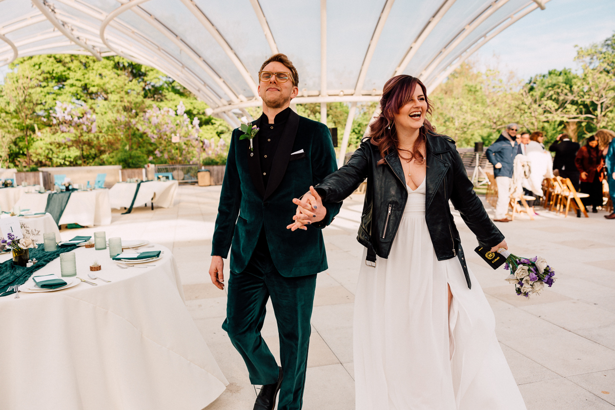 A stylish couple walks hand-in-hand, smiling happily, with wedding decorations and guests in the background under a beautiful glass canopy.