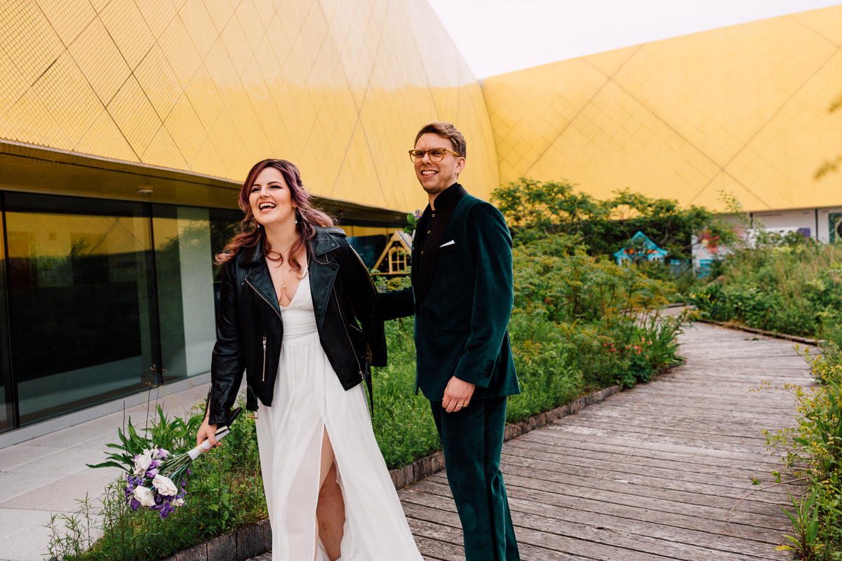 A bride in a leather jacket and a groom in a green suit share a laugh on a scenic wooden path surrounded by greenery and contemporary architecture.
