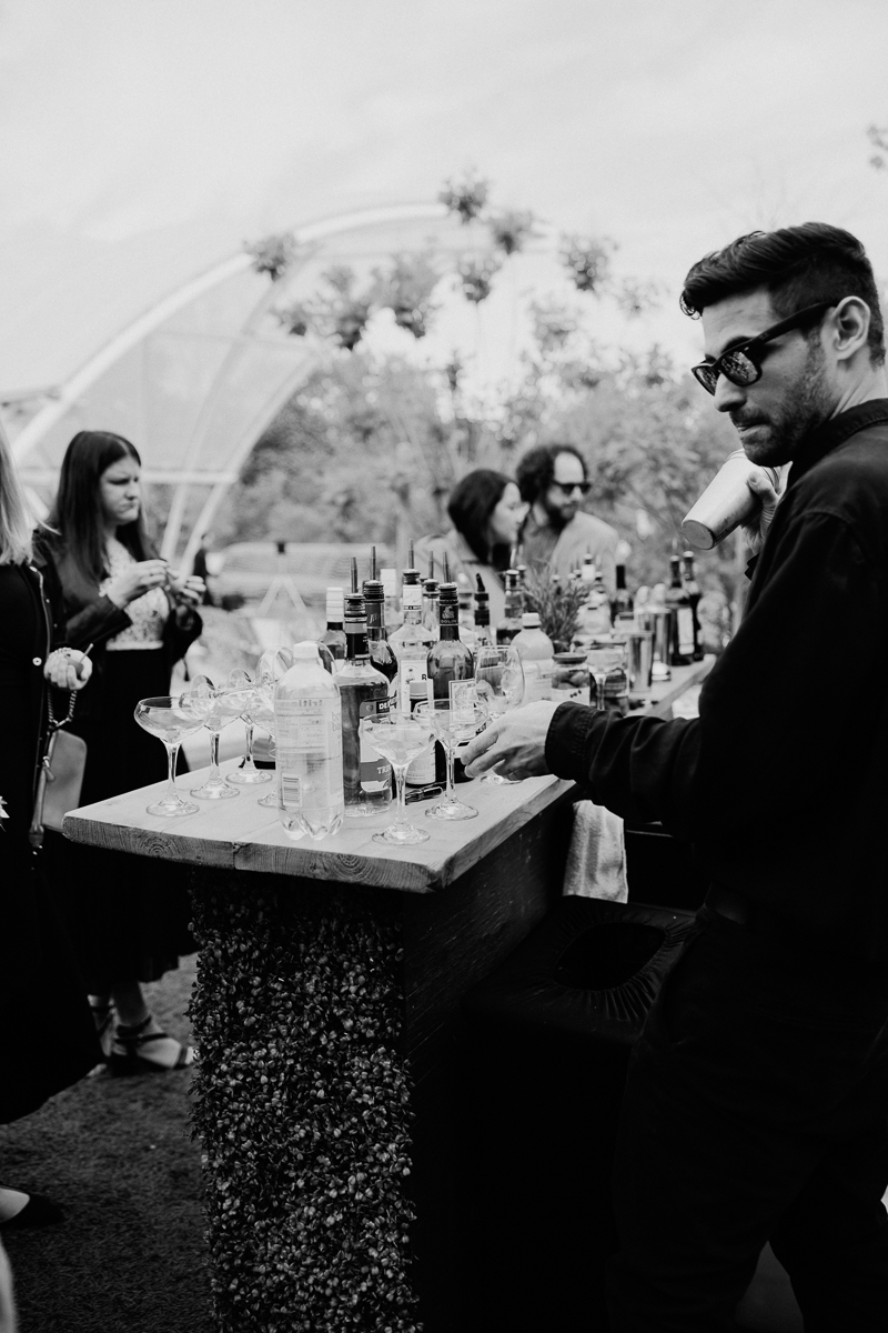 A bartender prepares drinks at a stylish outdoor bar, surrounded by guests enjoying the atmosphere, with a variety of bottles and glassware visible.