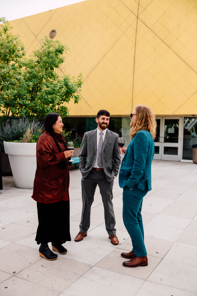 Three individuals in formal attire enjoying drinks outdoors in front of a modern yellow building.